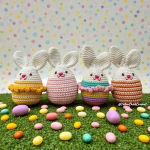 Puede incluir: Cuatro decoraciones de conejitos de Pascua hechas a mano con ganchillo. Cada conejito tiene cabeza y orejas blancas, nariz y boca bordadas en rosa y un cuerpo colorido. Los conejitos están sobre un lecho de césped artificial verde, rodeados de huevos de caramelo coloridos.