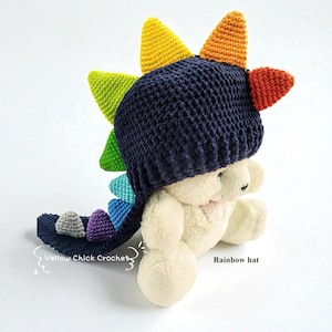 May include: A navy blue crochet hat with a rainbow-coloured dinosaur spine design. The hat is on a white stuffed animal. The hat has a tail with grey and purple accents. The text "Yellow Chick Crochet" and "Rainbow hat" are visible.
