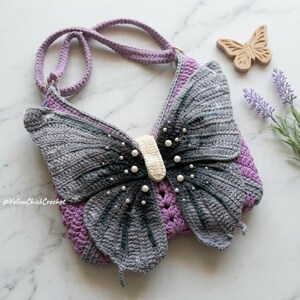 May include: A crochet shoulder bag shaped like a butterfly with grey and black gradient wings, a cream-colored center, and white pearl accents. The bag features a purple crocheted base and a matching purple strap, with the text YellowChickCrochet.