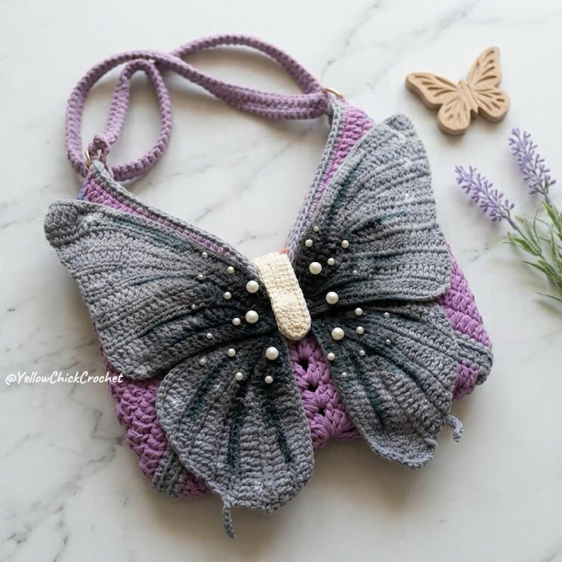 May include: A crochet shoulder bag shaped like a butterfly with grey and black gradient wings, a cream-colored center, and white pearl accents. The bag features a purple crocheted base and a matching purple strap, with the text YellowChickCrochet.
