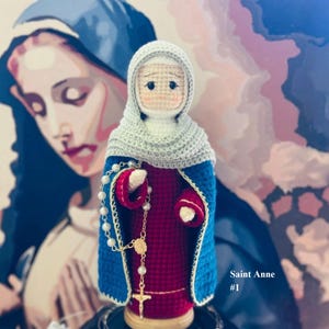 May include: A handmade crochet doll of Saint Anne, wearing a blue and red robe with a white head covering. A rosary with a gold cross hangs from her hands. The text "Saint Anne #1" is visible.