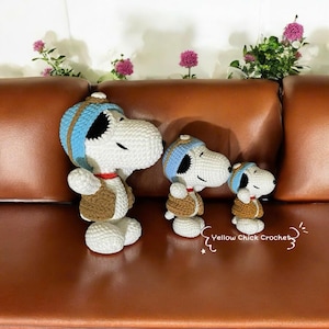 Può includere: Tre peluche di Snoopy, il personaggio dei cartoni animati, realizzati a uncinetto e con indosso gilet marroni e cappelli blu. I giocattoli sono disposti in fila, dal più grande al più piccolo, su un divano in pelle marrone. Il testo "Yellow Chick Crochet" è visibile nell'angolo in basso a destra dell'immagine.