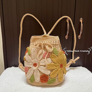 May include: A beige crochet backpack with a drawstring closure, adorned with large, colorful crochet flowers in cream, yellow, orange, pink, and green. The backpack has adjustable straps and the text "Yellow Chick Crochet".