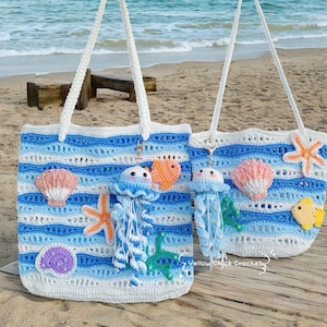 May include: Two crochet tote bags with white, light blue, and dark blue horizontal stripes. The bags are decorated with crochet sea creatures, including jellyfish, starfish, seashells, and fish. The bags have white handles and are on a sandy beach.