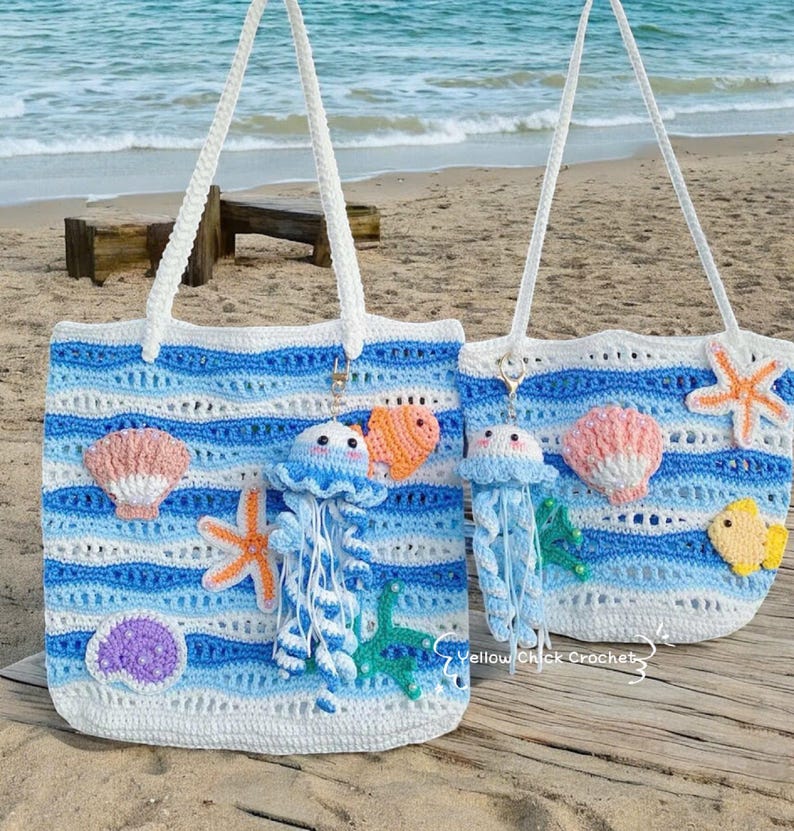 May include: Two crochet tote bags with white, light blue, and dark blue horizontal stripes. The bags are decorated with crochet sea creatures, including jellyfish, starfish, seashells, and fish. The bags have white handles and are on a sandy beach.