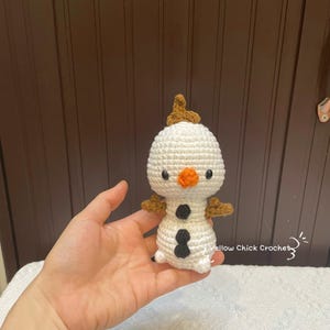 May include: A handmade crochet snowman doll with a white body, black button accents, and an orange carrot nose. The snowman has brown twig arms and a small brown hair tuft. The doll is held in a hand, with the text "Yellow Chick Crochet" visible.