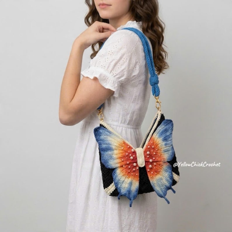 May include: A black crochet handbag featuring a large, three-dimensional butterfly design on the front. The butterfly wings are gradient-colored in orange, yellow, and blue with small white pearl accents. It has a thick blue crochet shoulder strap.