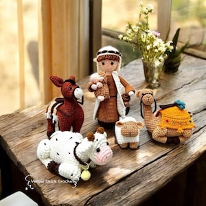 May include: A collection of handmade crochet nativity scene figures, including a shepherd, donkey, camel, cow, and sheep. The figures are in various colors, with detailed stitching and a rustic aesthetic. The scene is set on a wooden surface.