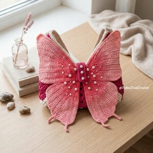May include: A crochet handbag shaped like a butterfly with pink and magenta yarn wings. The center of the butterfly features a vertical strip adorned with small white faux pearls. The bag has a textured, handmade appearance with visible stitching.