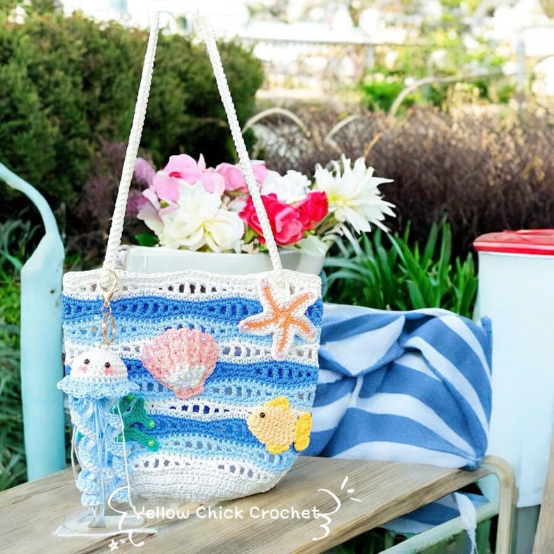 May include: A handmade crochet tote bag with a beach theme. The bag features blue and white stripes, and is decorated with a starfish, seashell, and fish. The bag has white rope handles and a jellyfish charm. The text "Yellow Chick Crochet" is visible.