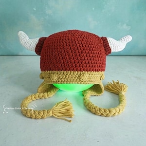 May include: A crocheted hat in deep red with a gold band, featuring two white horns on the sides and two long gold braided tassels hanging from the ear flaps. The text Yellow Chick Crochet is visible in the lower left corner of the image.