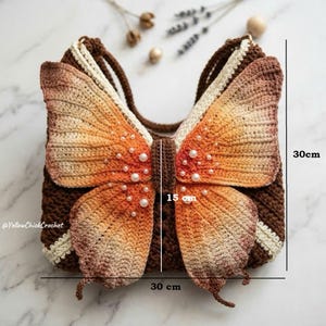 May include: A brown crochet bag shaped like a butterfly with orange and cream gradient wings, embellished with white faux pearls. The bag features a brown strap and measures 30 cm in height, 30 cm in width, and 15 cm in depth. Text reads YellowChickCrochet.