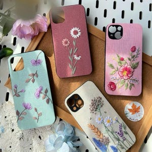 Puede incluir: Cuatro fundas de teléfono bordadas en varios colores. Una es verde claro con flores moradas, otra es rosa empolvado con margaritas blancas, una tercera es rosa con flores rojas y la última es blanquecina con un diseño floral.