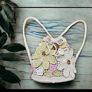 May include: A cream-coloured crochet backpack with a drawstring closure. The bag features large, three-dimensional crochet flowers in green, white, and pink. The straps are also crocheted. It is set against a light blue and green wooden background.