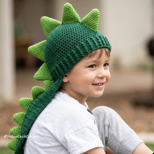 May include: A dark green crocheted hat featuring a series of light green triangular spikes running from the top down to the back. The hat has a ribbed brim and includes the text YellowChickCrochet printed in white on the lower left side.