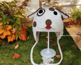 Pode incluir: Um chapéu branco de crochê em forma de cão dálmata, com manchas marrons, orelhas e uma língua vermelha. O chapéu tem longas amarrações trançadas com borlas. O chapéu é exibido em um suporte branco.