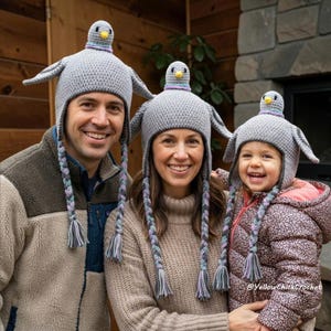 Puede incluir: Tres gorros de ganchillo grises a juego con un diseño de paloma en la parte superior, con orejeras y borlas trenzadas en tonos gris, azul y morado. Cada gorro muestra una pequeña cabeza de paloma rellena con un pico amarillo y cuello morado.