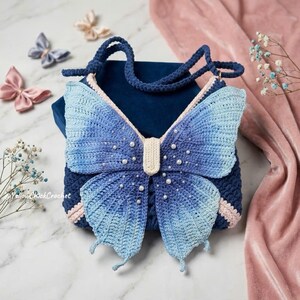 May include: A dark blue crochet bag featuring a large butterfly design on the front. The butterfly wings are ombre blue with white pearl accents. The bag has a cream-colored center body and a matching crochet strap. The text YellowChickCrochet is visible.