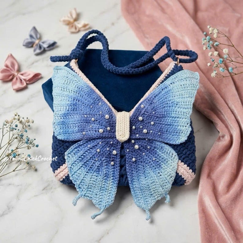 May include: A dark blue crochet bag featuring a large butterfly design on the front. The butterfly wings are ombre blue with white pearl accents. The bag has a cream-colored center body and a matching crochet strap. The text YellowChickCrochet is visible.