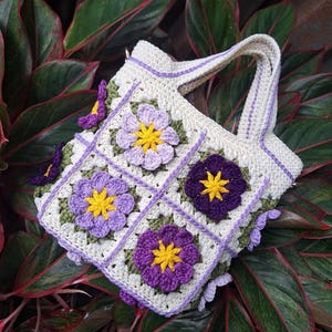 Op de afbeelding: Een handgemaakte gehaakte draagtas met een bloemenontwerp. De tas is voornamelijk crèmekleurig met paarse, gele en groene bloemenapplicaties. De tas heeft een stevige handgreep en staat tegen een achtergrond van groene en rode bladeren.