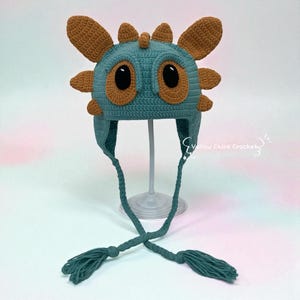 May include: A handmade crochet hat in teal and brown, featuring large, round eyes and ear-like projections. The hat has long ties with tassels. The text "Yellow Chick Crochet" is visible on the right side of the image. This whimsical hat is perfect for costume or everyday wear.