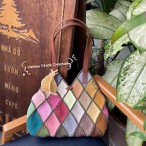 Peut inclure: Un sac fourre-tout en crochet coloré avec des anses en cuir marron. Le sac est composé de carrés de différentes couleurs, dont le rose, le vert, le bleu, le jaune et le marron. Le sac est posé sur une chaise en bois.
