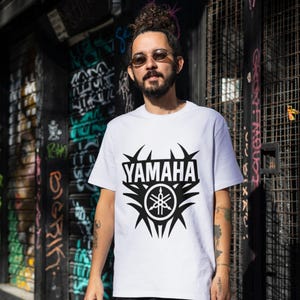 Pu&ograve; includere: T-shirt bianca con il logo Yamaha nero. Il logo include il nome del marchio e un emblema stilizzato. La maglietta a maniche corte &egrave; probabilmente in cotone.