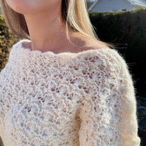 May include: Cream-coloured, off-the-shoulder crochet sweater. The sweater has a detailed, openwork stitch pattern, with a textured appearance. The garment is made of a soft yarn, possibly wool or cotton. The sweater is a light, neutral colour, suitable for various outfits.