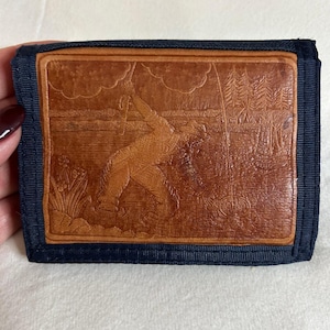 May include: A tri-fold wallet with a brown leather panel featuring an embossed scene of a person fishing. The wallet has a navy blue fabric border. The leather panel shows a person fishing in a lake with trees and clouds.