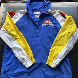 May include: A blue, yellow, and white Denver Nuggets hooded jacket. The jacket has a half-zip closure, a hood, and the Denver Nuggets logo on the chest. The sleeves are yellow and white with a rainbow accent.