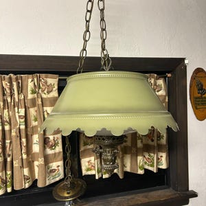 May include: A vintage hanging lamp with a pale green scalloped shade and ornate brass detailing. The lamp is suspended by a chain and has a decorative base. The lamp is positioned in front of a window with patterned curtains.