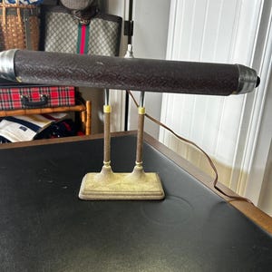 May include: A desk lamp featuring a long, horizontal, dark brown textured shade with silver-toned end caps. The lamp has two vertical support posts with brown textured accents and gold-toned rings, mounted on a rectangular, textured gold base.