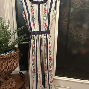 May include: A sleeveless dress with a cream background, adorned with vertical navy blue stripes and rows of red bows and floral patterns. The dress features a navy blue polka dot belt and trim.