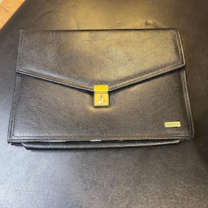 May include: Black leather briefcase with a gold-colored lock and the brand name "FUJITAKA" in gold lettering. The briefcase has a flap closure and a rectangular shape. The leather appears to be textured.