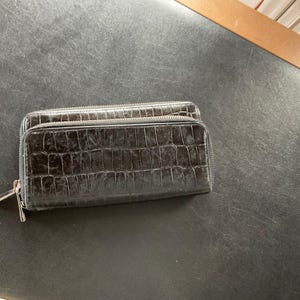 May include: A black, rectangular wallet with a textured, crocodile-like pattern. The wallet has two zippered compartments and a silver zipper pull. The wallet is made of a dark material.