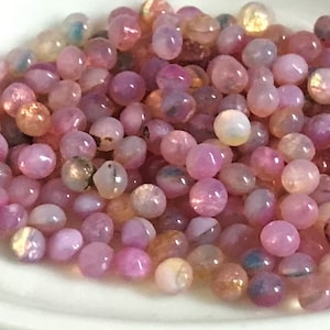 May include: A white ceramic spoon filled with small, iridescent pink and white beads. The beads have a pearlescent finish and are scattered randomly in the spoon.