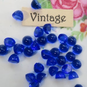 May include: A collection of vintage blue glass cabochons, also known as dome beads, on a white surface. The word "Vintage" is written on a small piece of paper.