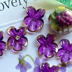 May include: Purple glass flower buttons with gold trim. The buttons are arranged on a white surface with a floral pattern.