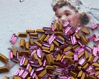 May include: A large pile of pink and gold glass rhinestones. The rhinestones are in various shapes and sizes, including rectangular, square, and round.
