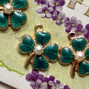 Puede incluir: Tres colgantes de tr&eacute;bol de cuatro hojas verdes y dorados con un centro de perla. Los colgantes est&aacute;n colocados sobre un fondo floral vintage con flores moradas y la palabra "FR" visible. Los colgantes tienen un lazo dorado para colgar.
