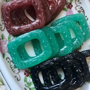 May include: Assortment of vintage plastic buckles in various colors, including black, brown, green, and red. The buckles are displayed on a white porcelain dish with floral designs.