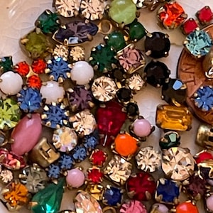 May include: A collection of colorful rhinestones and small jewels in various shapes and sizes. The gems are in shades of red, blue, green, pink, and yellow, with gold-colored metal settings. A penny is included for size reference.
