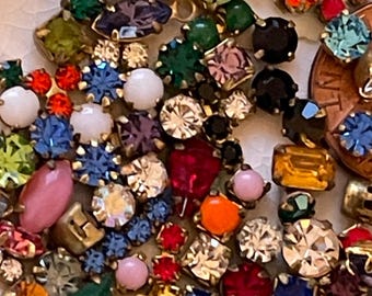 Könnte beinhalten: Eine Sammlung bunter Strasssteine und kleiner Juwelen in verschiedenen Formen und Größen. Die Edelsteine sind in den Farbtönen Rot, Blau, Grün, Rosa und Gelb gehalten, mit goldfarbenen Metalleinfassungen. Eine Münze dient als Größenreferenz.