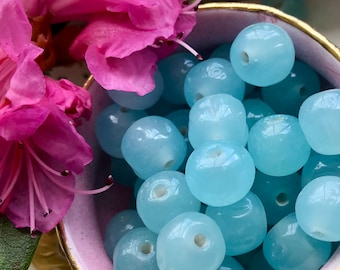 May include: A bowl filled with light blue glass beads. The beads are round and have a smooth, polished finish. The bowl is pink with a gold rim.