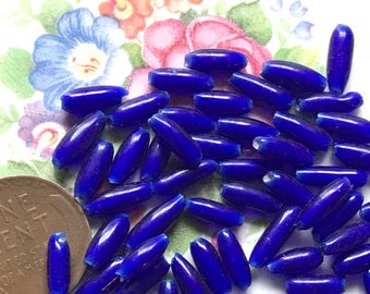 May include: A pile of cobalt blue glass beads, each bead is oval shaped and about 2.5 cm long. The beads are scattered on a white surface.