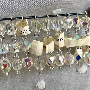 May include: A strand of iridescent crystal beads hangs from a silver wire. The beads are faceted and have a rainbow sheen. A torn piece of paper with the text "MADE IN JAPAN" is attached to the wire.