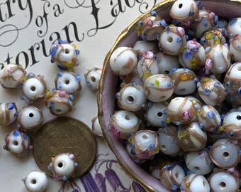 May include: A bowl filled with white glass beads with gold and blue accents. The beads have a variety of patterns and designs.
