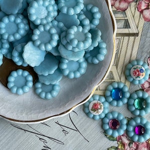 May include: A collection of light blue glass cabochons with a beaded edge, displayed in a white dish with a gold rim. There are also several cabochons with floral and jewel accents.