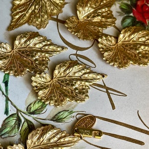 Peut inclure: Un ensemble de breloques en forme de feuille en métal doré avec une surface texturée détaillée. Les breloques ont la forme d'une feuille avec une pointe pointue et une base arrondie. Les breloques sont éparpillées sur une surface blanche avec un motif floral.