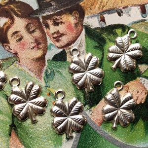 May include: Six silver four-leaf clover charms with a loop at the top for attaching to a necklace or bracelet. The charms are on a green and white background.